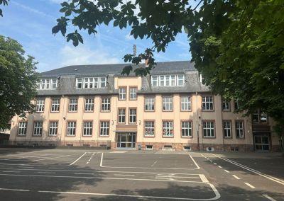 Blick auf ein großes, helles Schulgebäude mit mehreren Etagen und einem leeren, markierten Schulhof im Vordergrund. Bäume rahmen das Bild von oben und an den Seiten.