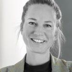 Frau mit hochgesteckten blonden Haaren, trägt helles Jackett und dunkles Oberteil, vor grauem Hintergrund
