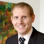 Joachim Sittig – Zentrum für Weiterbildung Joachim Sittig - Porträt eines Mannes in Anzug und gestreifter Krawatte. Er hat kurzes blondes Haar und lächelt freundlich in die Kamera. Im Hintergrund ist ein buntes Gemälde zu sehen.