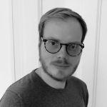 Portrait von Marc Kämpf - Mann mit Brille, Bart und kurzem Haar, trägt ein dunkles T-Shirt und steht vor einer weißen Wand