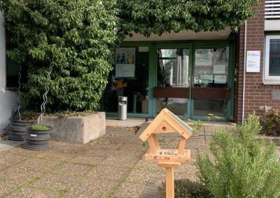 Zentrum für Weiterbildung – Standort Rüsselsheim Eingangsbereich des Zentrums für Weiterbildung in Rüsselsheim mit begrüntem Vorplatz und einem Holz-Vogelhäuschen im Vordergrund.