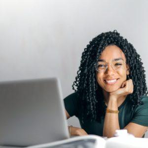 Junge Frau mit Laptop lächelt direkt in die Kamera, ihr Kopf ist aufgestützt, sie sitzt vor weißem Hintergrund.