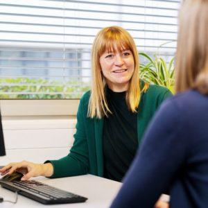Frau führt ein Beratungsgespräch an einem Schreibtisch, im Vordergrund sitzt eine andere Person.