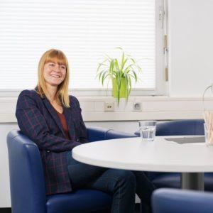 Frau sitzt in modernem Beratungszimmer entspannt an einem runden Tisch und lächelt.