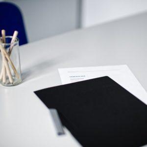 Bewerbungsunterlagen und Stift auf einem Tisch, im Hintergrund ein Glas mit Holzstiften.