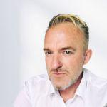 Marc Kämpf – Zentrum für Weiterbildung Denis Götz - Porträt eines Mannes mit kurz geschnittenem blonden Haar, leichtem Bart und weißem Hemd vor hellem Hintergrund.