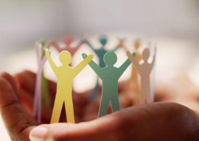 Zwei Hände halten eine Gruppe aus bunten Papierfiguren, die sich an den Händen halten und einen Kreis bilden.