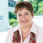 Pia Moser-Manthei – Zentrum für Weiterbildung Portrait Pia Moser-Manthei - einer Frau mit kurzen, braunen, gelockten Haaren. Sie trägt ein weißes Hemd und einen rotgemusterten Schal und steht vor einem Fenster.