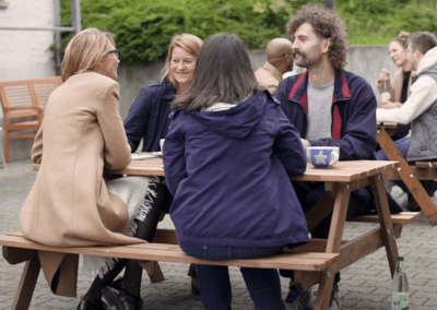 Vier Personen sitzen in einer kleinen Gruppe an einem Picknicktisch im Freien und unterhalten sich freundlich. Weitere Menschen sitzen im Hintergrund an anderen Tischen.