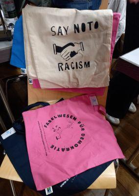 Zwei bedruckte Stoffbeutel auf einem Stuhl mit Aufschriften gegen Rassismus und für Demokratie.