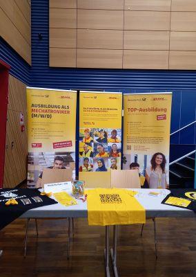Gelber Infostand von DHL mit Roll-Ups zu Ausbildungsplätzen und Werbegeschenken auf dem Tisch.