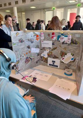 Eine junge Frau mit Kopfhörern sitzt vor einem kreativ gestalteten Sichtschutz aus Zeitungen und Bildern. Im Hintergrund stehen mehrere Jugendliche in einer Messehalle.