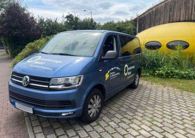 Blaues Beratungsfahrzeug mit der Aufschrift „Orientierungsmobil“ steht auf einem gepflasterten Parkplatz vor einem gelben, rundlichen Gebäude.