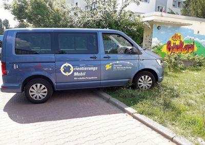 Ein blauer Kleinbus mit der Aufschrift „OrientierungsMobil – Mit unschlagbarer Perspektive“ steht am Straßenrand neben einer bunten Wand mit dem Schriftzug „die Bäckerei“.