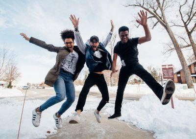 Drei junge Menschen springen lachend in die Luft auf einem verschneiten Gehweg – voller Energie und Freude.