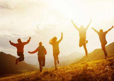 Vier Personen springen mit erhobenen Armen auf einer Wiese vor einer Berglandschaft im warmen Sonnenlicht.