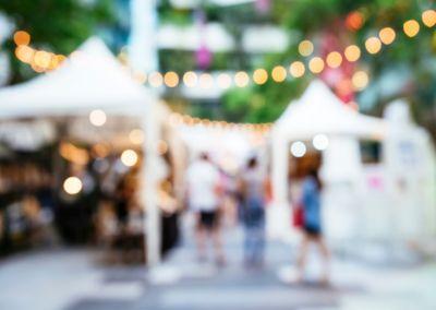 Unscharfes Foto einer Außenveranstaltung mit weißen Pavillonzelten. Zwischen den Zelten hängen Lichterketten mit warmem, gelbem Licht. Mehrere Personen bewegen sich im Vordergrund über einen gepflasterten Platz. Im Hintergrund sind grüne Bäume und Gebäude erkennbar. Die Atmosphäre wirkt festlich und sommerlich.