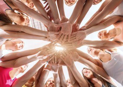 Mehrere Personen stehen im Kreis und strecken ihre Hände in die Mitte, wo sie sich übereinanderlegen; von unten fotografiert mit Blick in einen hellen Himmel.