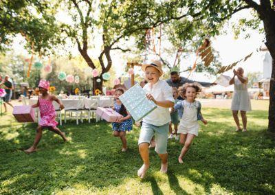 Mehrere Kinder rennen fröhlich über eine Wiese bei einer Gartenfeier und tragen Geschenke, während Erwachsene im Hintergrund klatschen und dekorierte Tische unter Bäumen stehen.