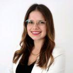 Sally Kiwus aus der Unternehmenskommunikation lächelt mit Brille und weißem Blazer vor hellem Hintergrund in die Kamera.